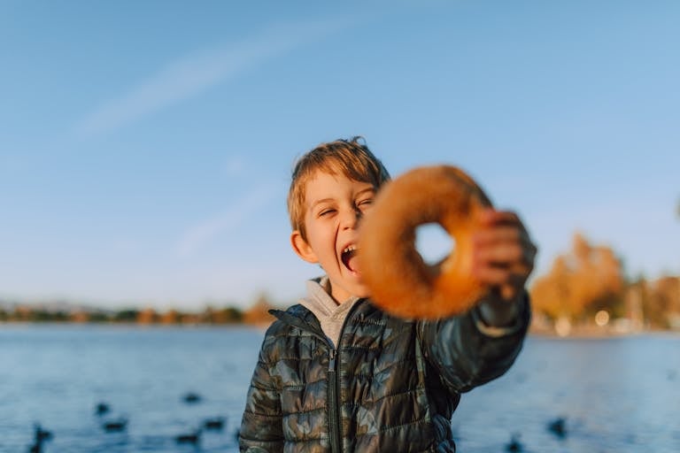 Happy child holding a bagel at a lakeside, enjoying a sunny outdoor snack.